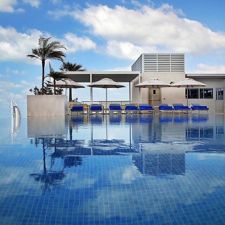 Grand Cosmopolitan Hotel Dubai Luaran gambar The photo depicts a luxurious swimming pool with a stunning infinity edge. The water is calm and reflects the sky, creating a serene atmosphere. Surrounding the pool are several blue lounge chairs with white umbrellas, providing spots for relaxation.