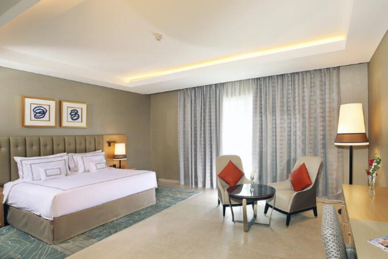Grand Cosmopolitan Hotel Dubai Luaran gambar The photo shows a modern hotel room featuring a spacious layout. There is a large bed with a headboard adorned with decorative artwork above it. On one side of the bed, there's a bedside lamp. Opposite the bed, there are two comfortable armchairs sty