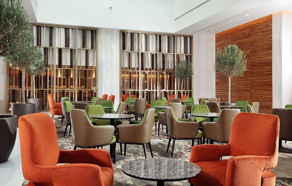 Grand Cosmopolitan Hotel Dubai Restoran gambar The photo shows a modern, stylish lounge area with comfortable seating. The space features multiple tables surrounded by chairs in vibrant colors, including orange and green. There are also decorative plants in the corners, adding a touch of greenery