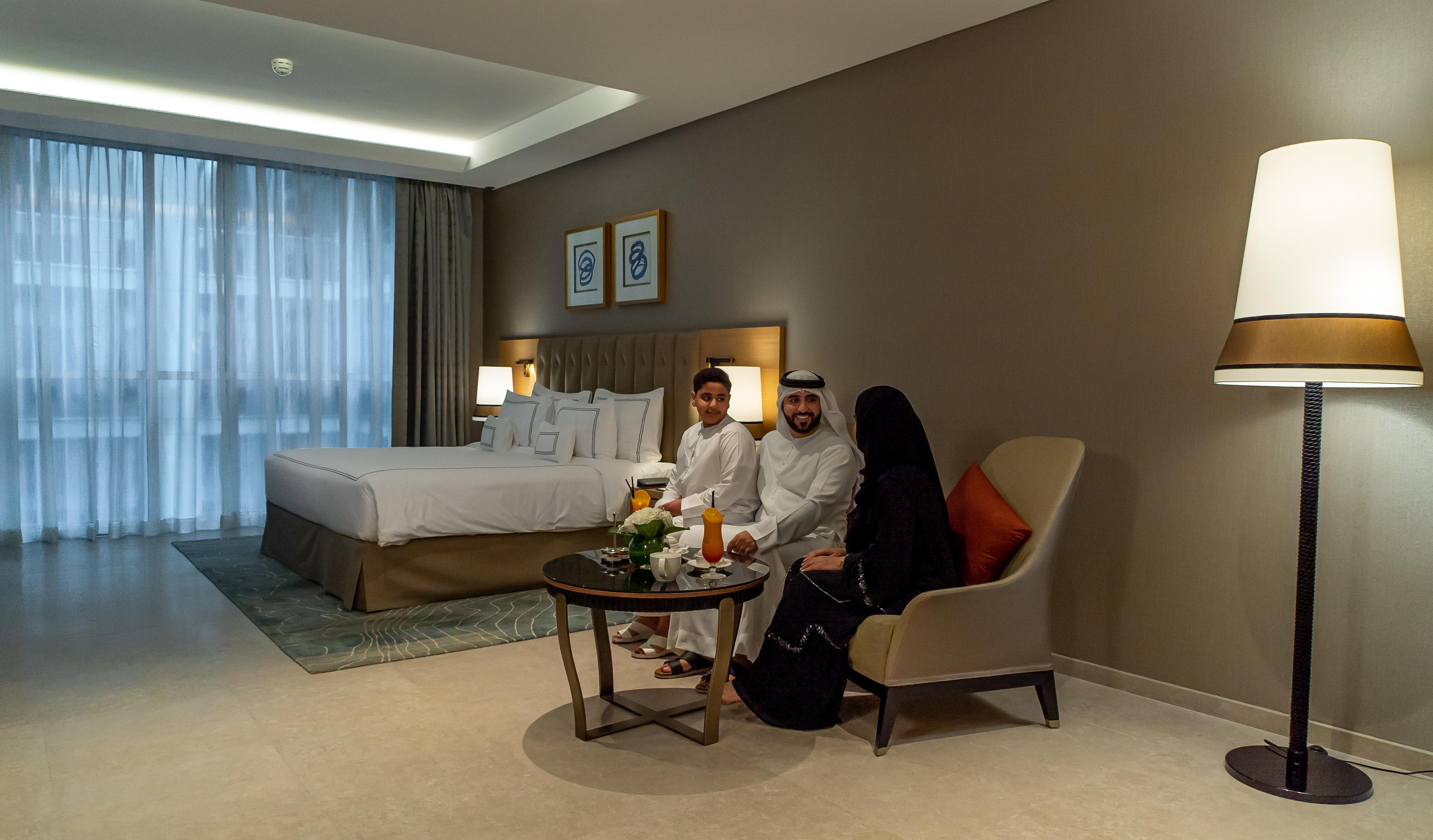 Grand Cosmopolitan Hotel Dubai Luaran gambar The photo shows a modern hotel room featuring a large bed with white linens and a stylish headboard. In the room, there is a small round table with a floral centerpiece and drinks. Three individuals are seated on the chairs by the table: two adults a