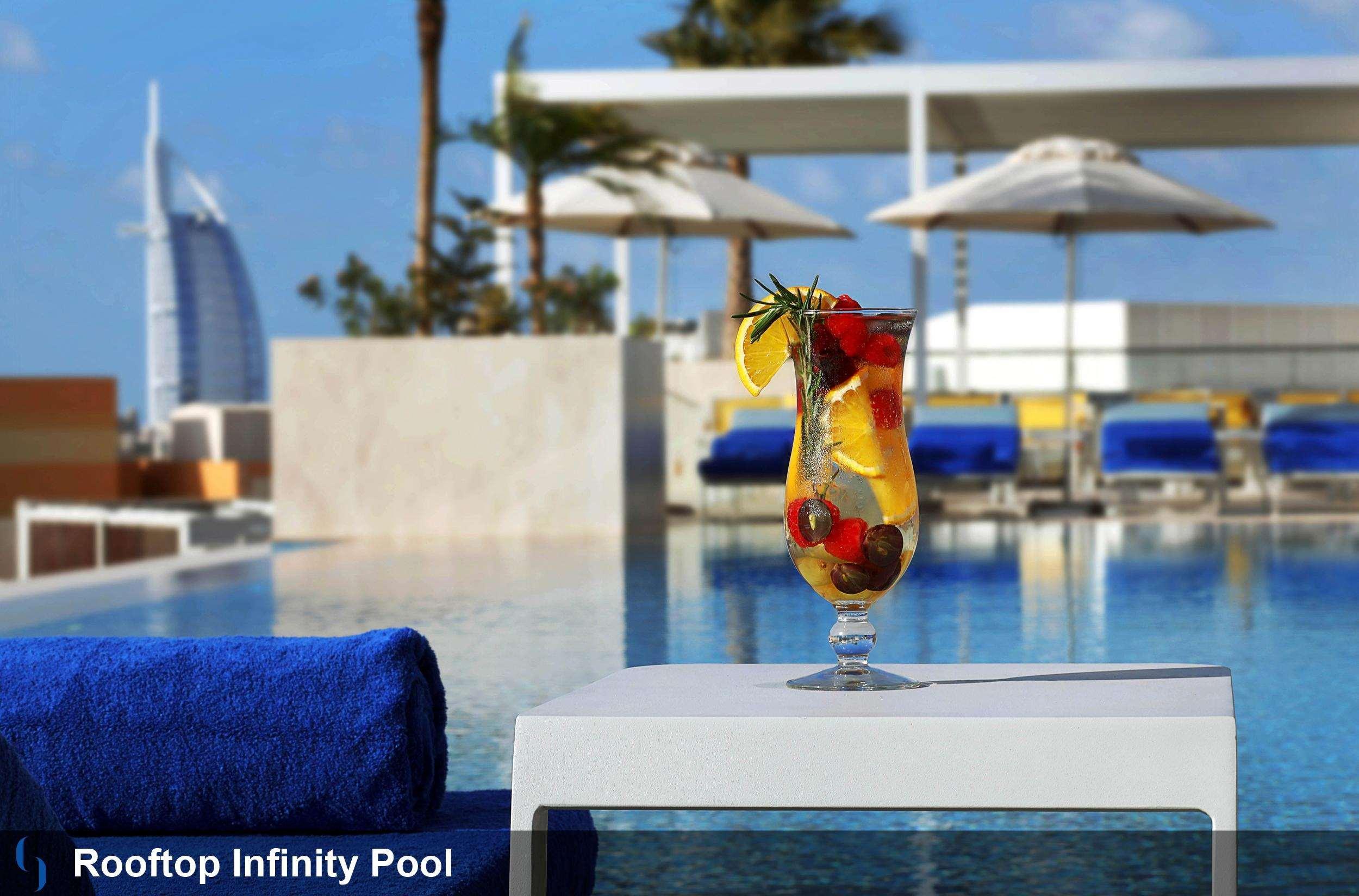 Grand Cosmopolitan Hotel Dubai Luaran gambar The photo shows a rooftop infinity pool with a stunning view. In the foreground, there is a cocktail glass filled with a colorful drink garnished with fruits, including lemon and berries. The background features cozy loungers and umbrellas, suggestin