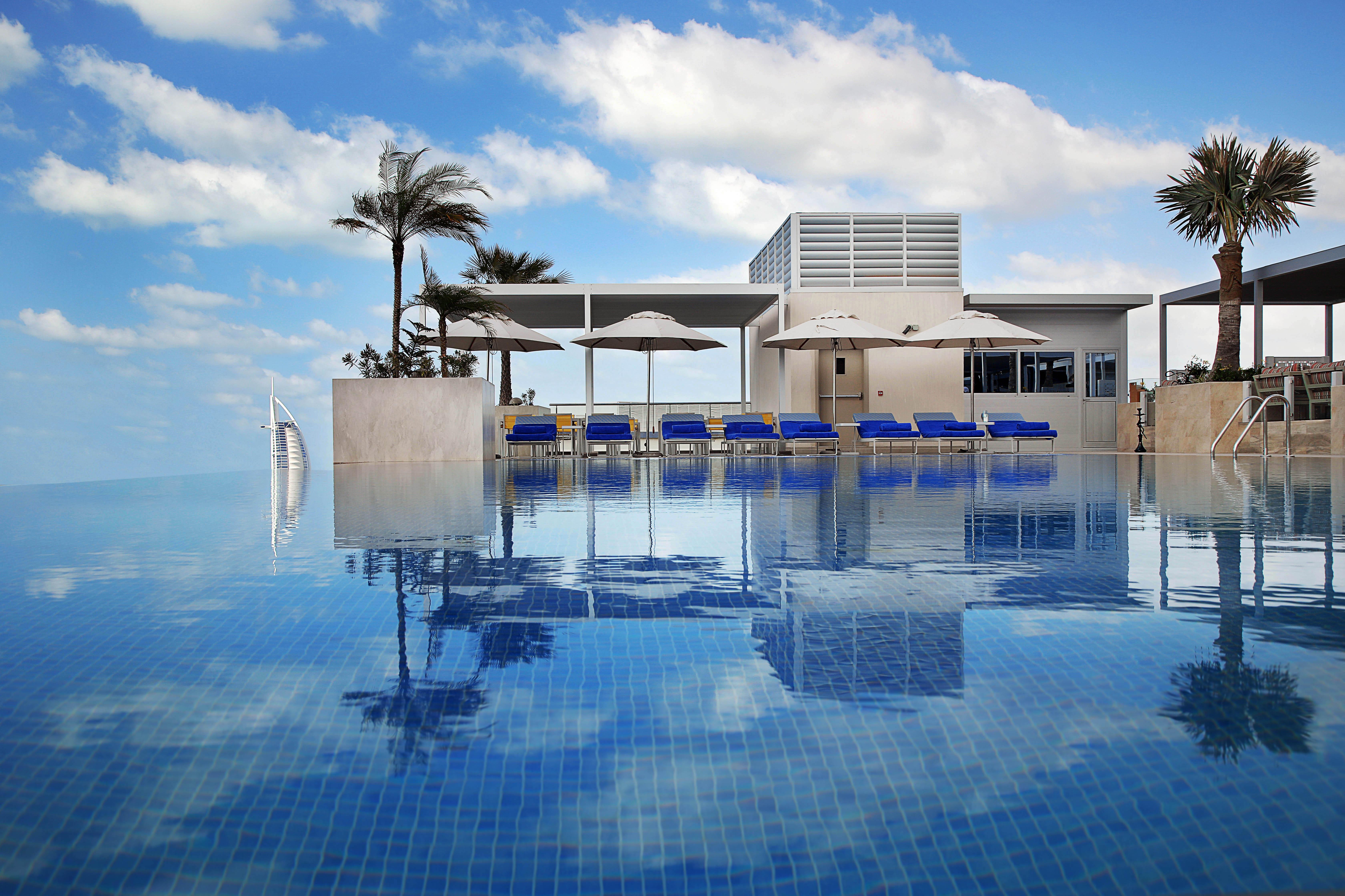 Grand Cosmopolitan Hotel Dubai Luaran gambar The photo depicts a luxurious swimming pool with a stunning infinity edge. The water is calm and reflects the sky, creating a serene atmosphere. Surrounding the pool are several blue lounge chairs with white umbrellas, providing spots for relaxation.