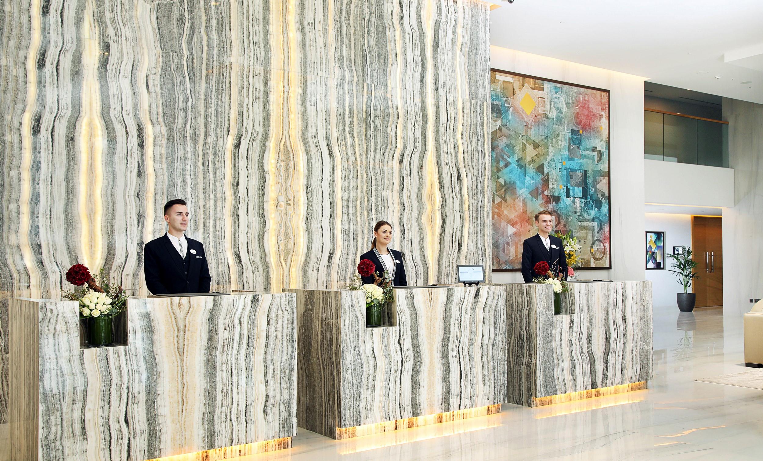 Grand Cosmopolitan Hotel Dubai Luaran gambar The photo shows a hotel reception area featuring three receptionists standing behind a sleek, modern front desk made of a striped marble-like material with illuminated edges. They are dressed in formal uniforms. Behind them, there is a large piece of