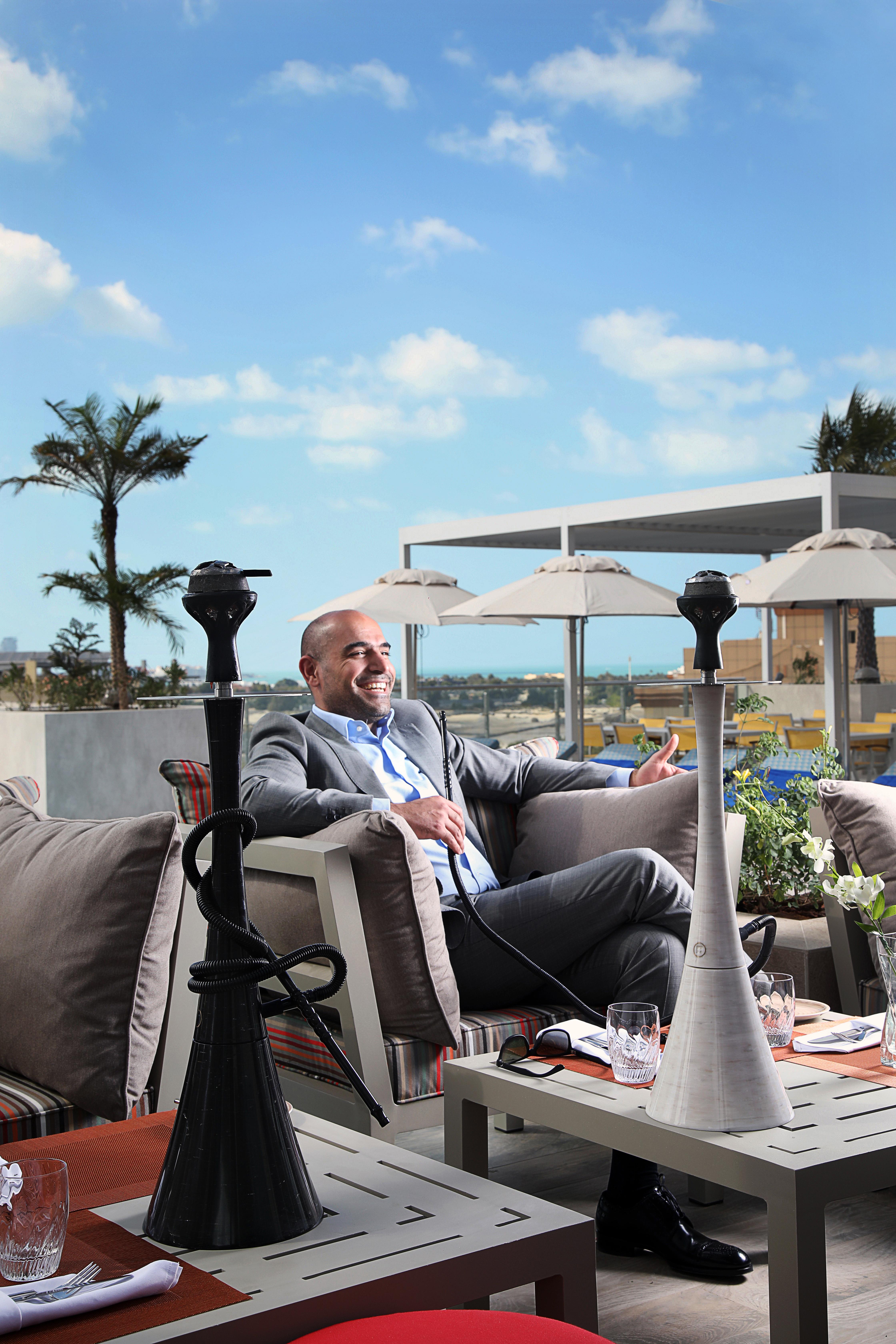 Grand Cosmopolitan Hotel Dubai Luaran gambar The image shows a man sitting comfortably on a sofa in an outdoor lounge area. He is dressed in a suit and is smiling, suggesting a relaxed atmosphere. In front of him are two hookahs, which are traditional smoking devices. The background features pa