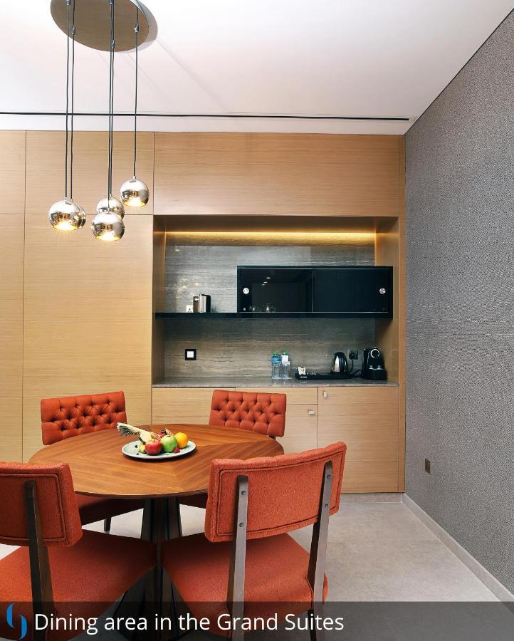 Grand Cosmopolitan Hotel Dubai Luaran gambar The photo depicts a modern kitchen area. There is a round dining table with orange upholstered chairs surrounding it. On the table, a bowl of assorted fruits is displayed. In the background, a countertop with some kitchen appliances, including a kett