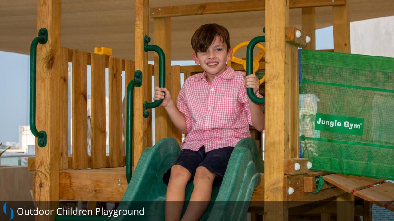 Grand Cosmopolitan Hotel Dubai Luaran gambar The image shows a young boy sitting on a slide in a children's playground. He is wearing a pink checkered shirt and dark shorts. The playground equipment is made of wood and features green sliding elements. The boy has a joyful expression, suggesting