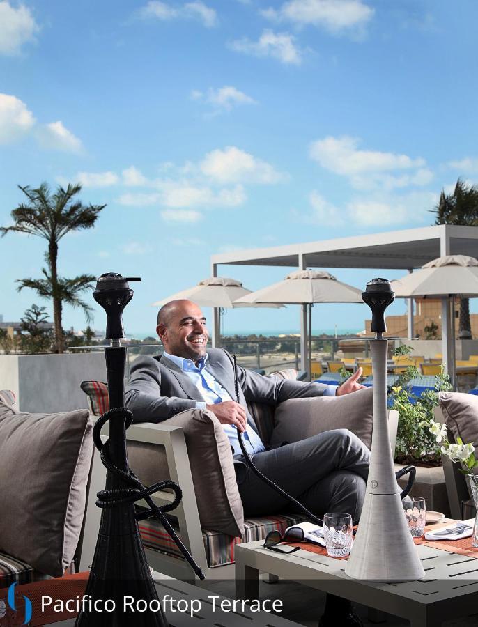 Grand Cosmopolitan Hotel Dubai Luaran gambar The photo shows a man sitting comfortably in an outdoor lounge area. He is dressed in a light grey suit and appears to be smiling and relaxed. In the background, there are several palm trees and sun loungers with umbrellas, suggesting a tropical or u