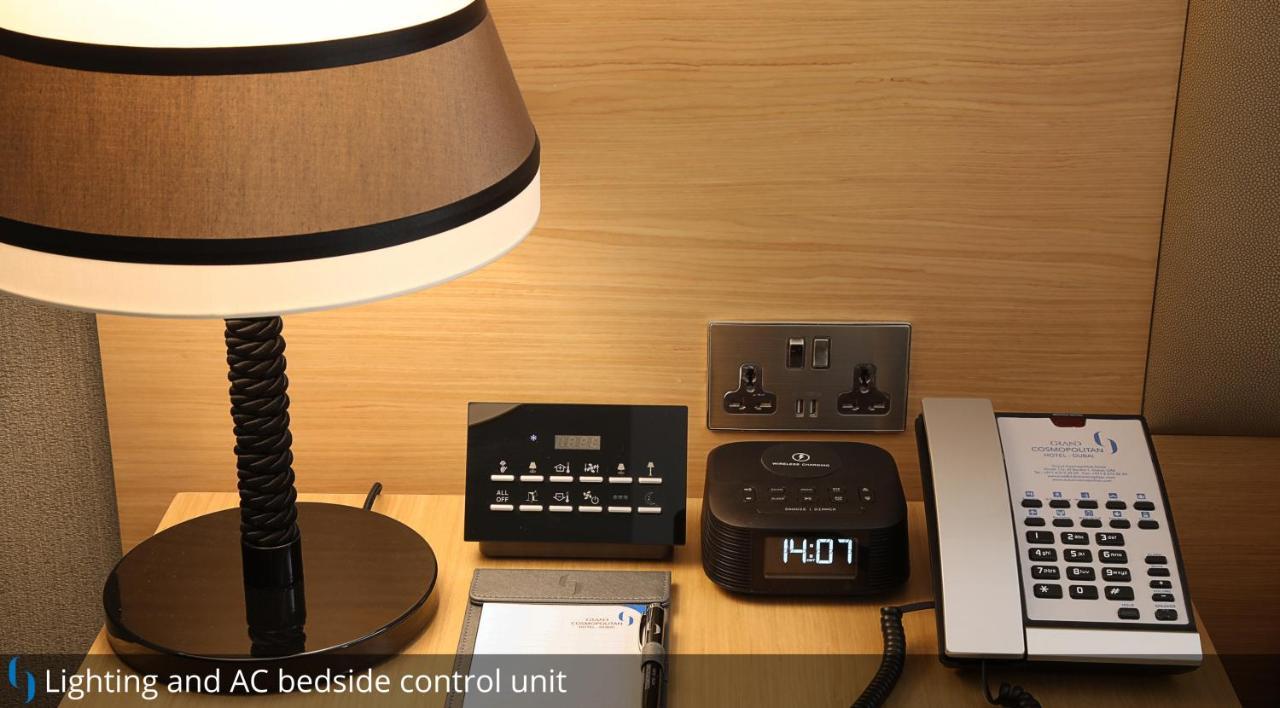 Grand Cosmopolitan Hotel Dubai Luaran gambar The photo shows a bedside setup with several items. There is a table lamp with a dark lampshade on the left. Next to it is a control unit with various buttons, likely for managing room settings such as lighting and air conditioning. Beside the contro