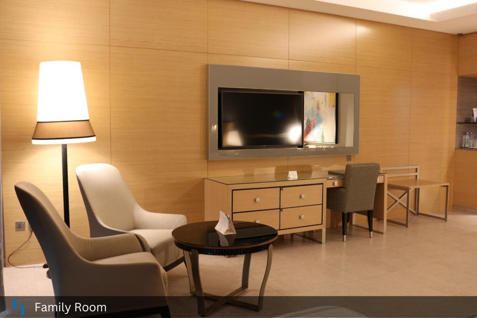 Grand Cosmopolitan Hotel Dubai Luaran gambar The photo shows a modern family room with a warm and inviting atmosphere. It features a light wood-paneled wall and contains several pieces of furniture:

- A flat-screen television mounted on the wall, positioned above a wooden media console with dr