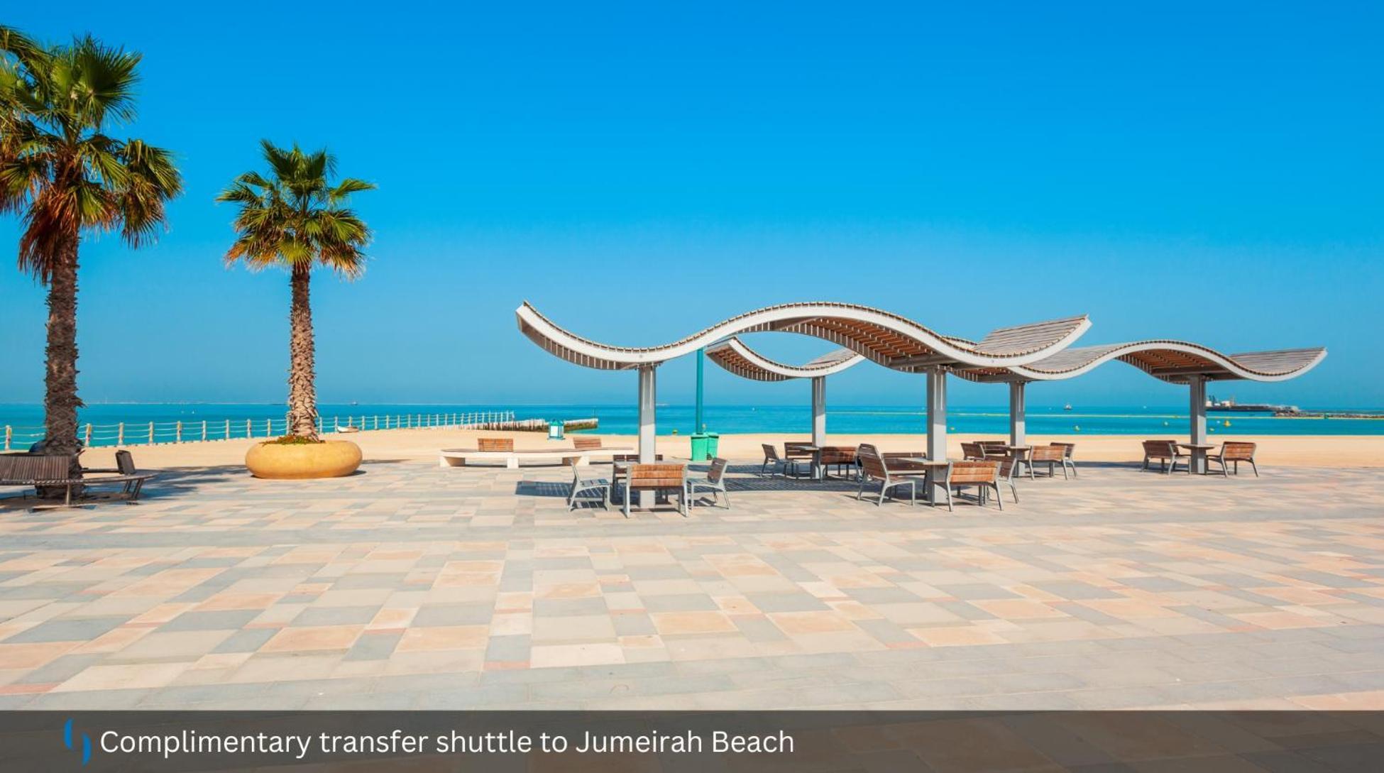 Grand Cosmopolitan Hotel Dubai Luaran gambar The photo shows a scenic beach area, likely at Jumeirah Beach. In the foreground, there are wooden benches and tables arranged under shaded structures with a wavy design. A tall palm tree adds to the tropical feel of the setting. In the background, y